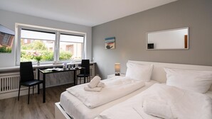 A modern hotel room with a bed, a desk, and a chair. There is a window with a view of buildings and greenery outside. - Adrian 3 - Gemütliches, Zentrales Apartment für 2 Personen, Wlan und TV, Separate Küche (Sylt)