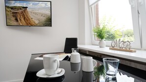 A modern room with a dining table, a coffee setup, a framed coastal landscape photo, a window with a plant, and a decorative word sign. - Adrian 3 - Gemütliches, Zentrales Apartment für 2 Personen, Wlan und TV, Separate Küche (Sylt)