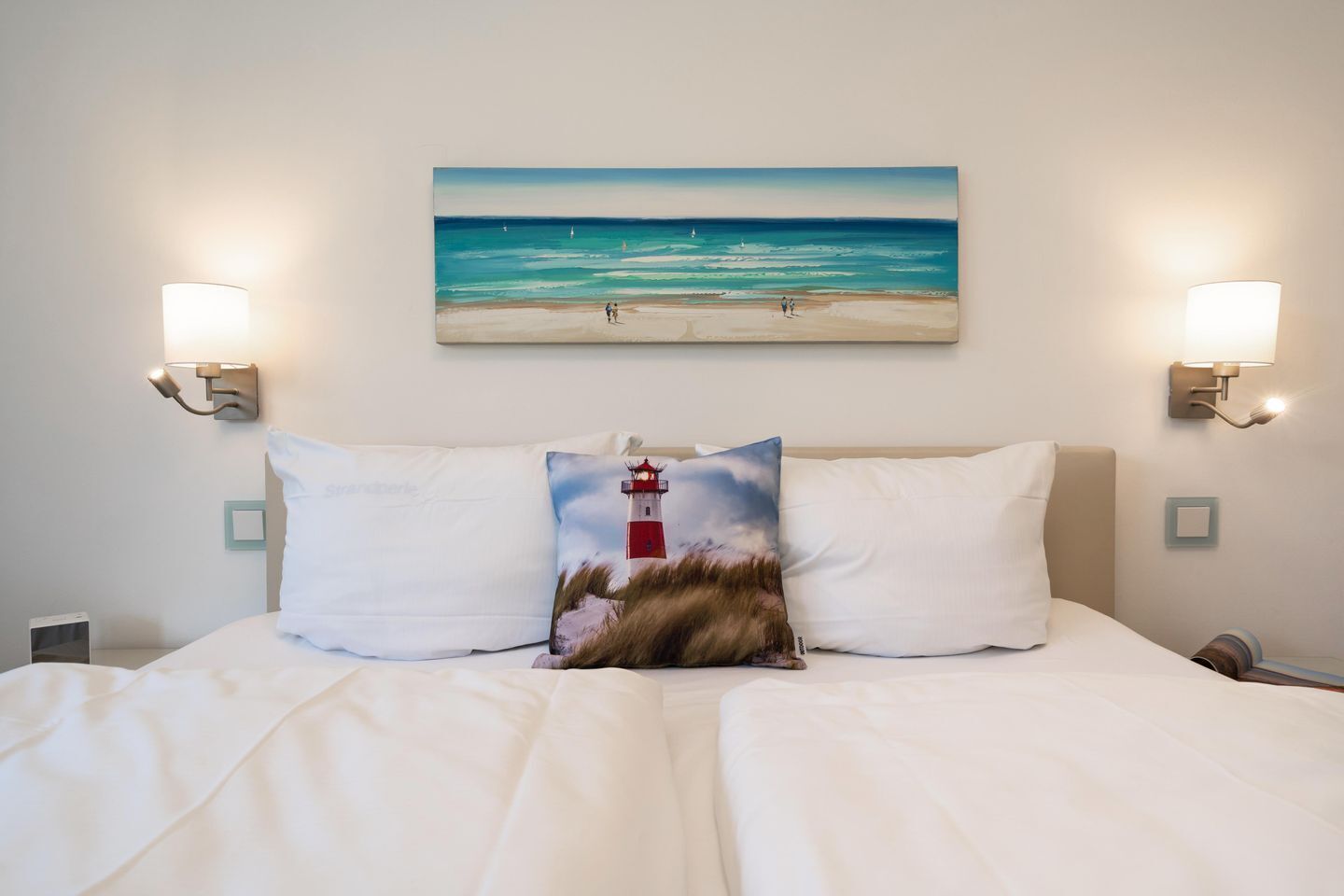 Habitación de hotel con cama, dos almohadas, un cuadro de una playa y un faro, y luces fijadas a la pared.