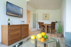 Una sala moderna con un aparador de madera, una mesa de vidrio con frutas, un comedor con mesa y sillas de madera, un cuadro en la pared y una planta.