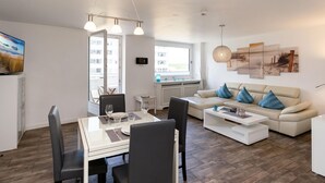En moderne stue med spiseplads, en sofa med blå puder og et stort vindue der lukker naturligt lys ind.