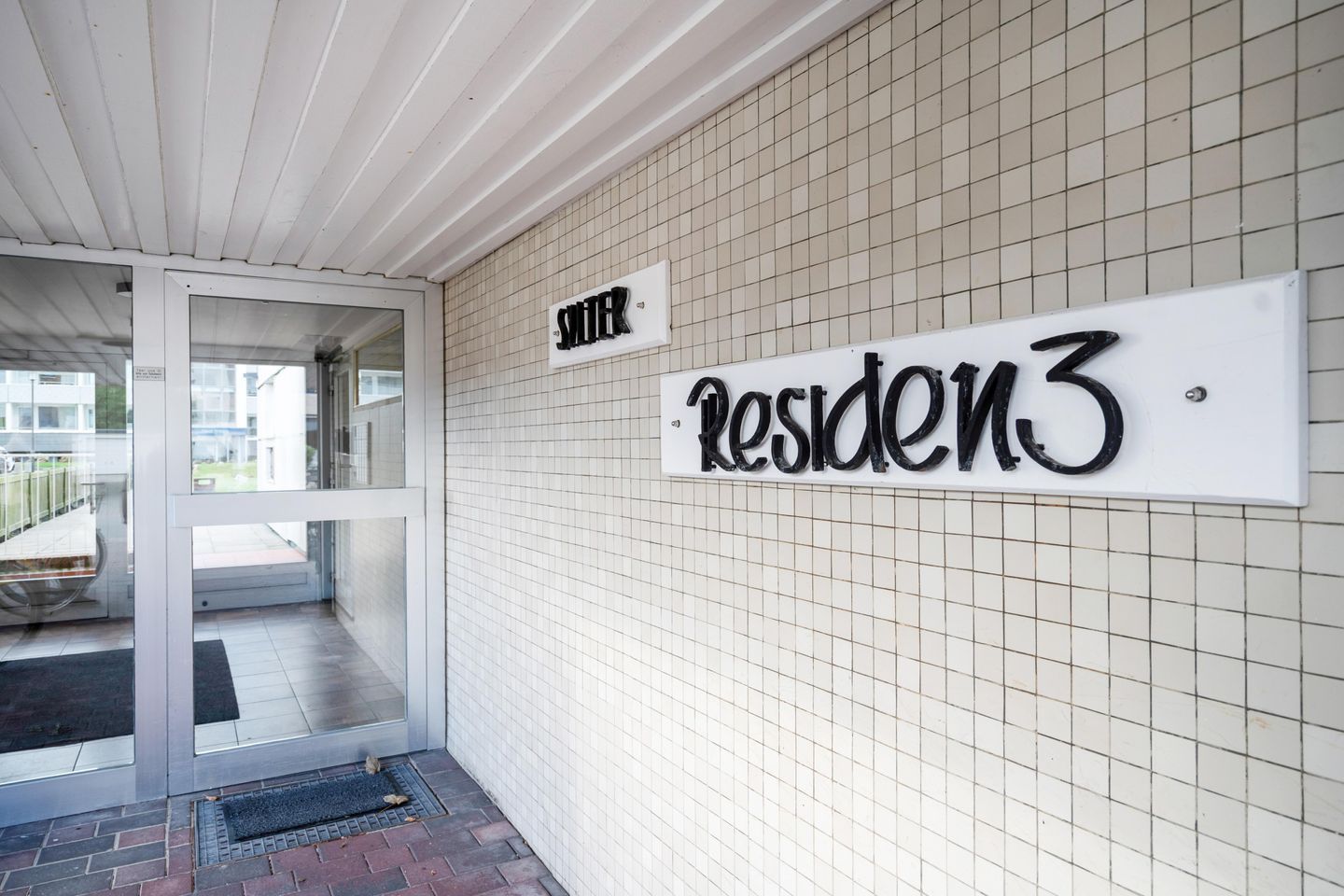 A building entrance with a glass door, a sign reading 'SITZER Residence 3', and a tiled wall.