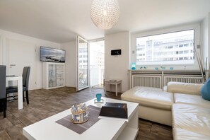 A modern living room with a white sofa, a coffee table, and a TV. There's a balcony with a view of buildings. - Sylter Residenz - Modern apartment near the beach, with a balcony, parking space, and Wi-Fi. (Sylt)