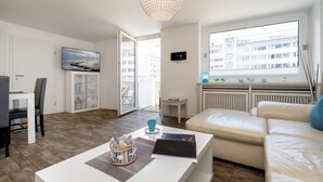 En moderne stue med en hvid sofa, et sofabord og et fjernsyn. Der er en balkon med udsigt til bygninger.