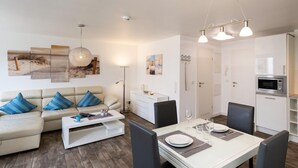 En moderne stue med sofa, sofabord og spisebord dækket til to personer. Der er et køkkenområde med mikroovn og skabe.