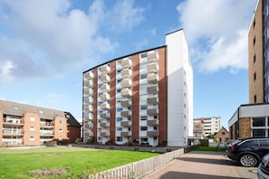 A multi-story residential building with balconies, a paved driveway, and a grassy area in front.