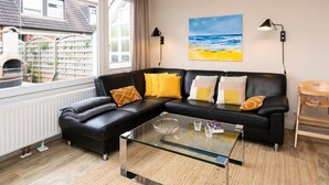A modern living room with a black sectional sofa, a glass coffee table, and a painting of a beach scene. - Süderliebe - Modern house for 6 pers. with terrace, parking space, WLAN. (Sylt)
