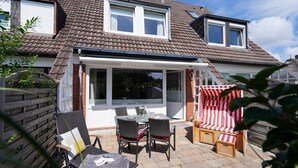 A two-story house with a patio area featuring a table and chairs, a red and white striped chair, and a wooden fence. - Süderliebe - Modern house for 6 pers. with terrace, parking space, WLAN. (Sylt)