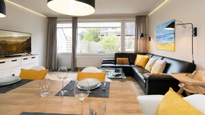 A modern living room with a black sofa, yellow cushions, a dining table set with plates and wine glasses, a TV on a cabinet, and a large window with a view of buildings and trees. - Süderliebe - Modern house for 6 pers. with terrace, parking space, WLAN. (Sylt)