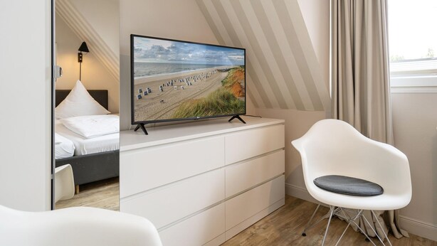 A modern hotel room with a bed, a flat-screen TV on a dresser, a white chair, and a view of the beach through the window. - Süderliebe - Modern house for 6 pers. with terrace, parking space, WLAN. (Sylt)