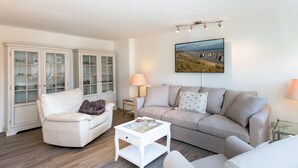 Un salón con un sofá blanco, un sillón blanco, una mesa de centro de vidrio y un aparador con puertas de cristal.