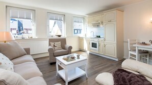Una sala moderna con sofá, sillón y mesa de centro. Hay una zona de cocina con armarios y estufa.