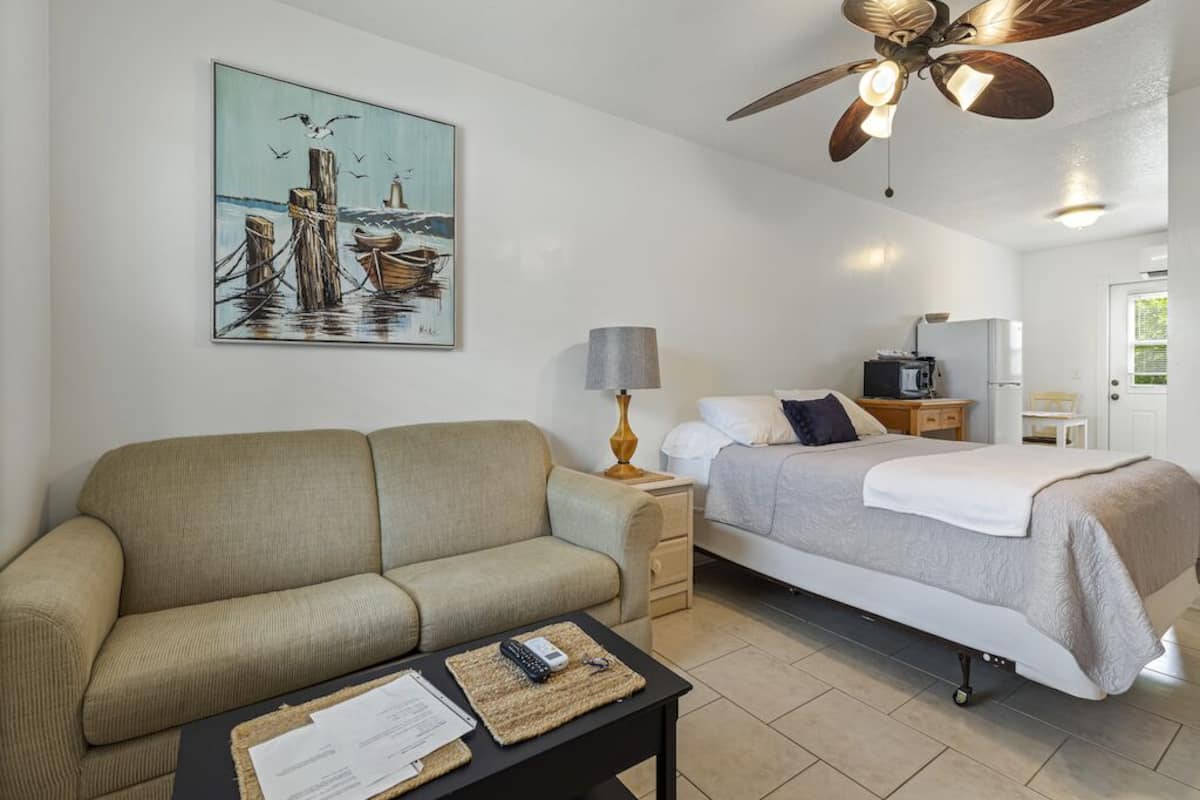 A hotel room with a bed, a sofa, a desk with a lamp, a ceiling fan, and a painting of a seascape.