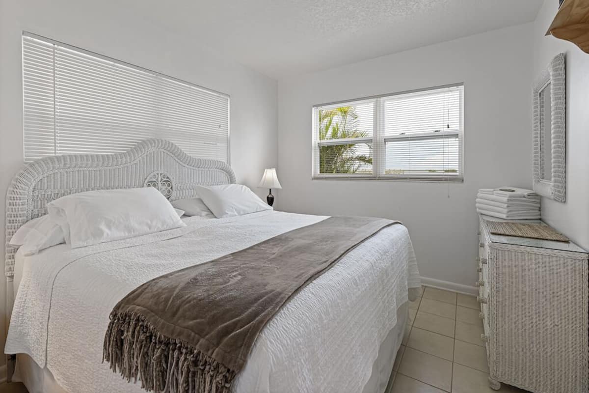 A bedroom with a large bed, white bedding, a grey throw blanket, a bedside table with a lamp, and a wicker side table with folded towels.