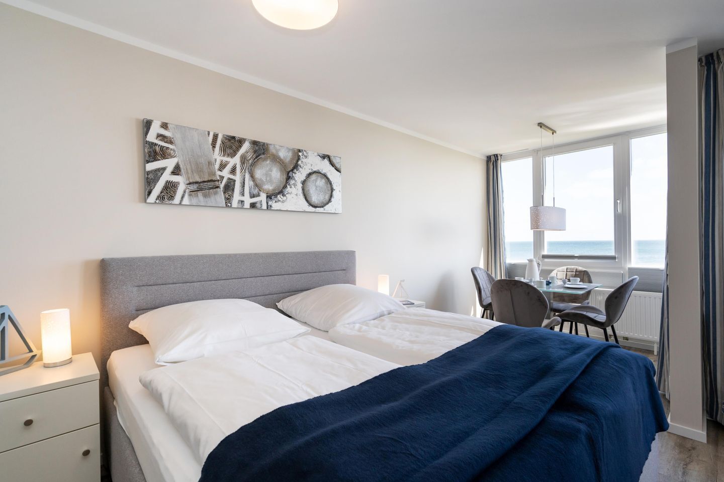 Een hotelkamer met een bed, nachtkastje, lamp en uitzicht op zee door grote ramen.