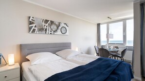 Habitación de hotel con cama, mesita de noche, lámpara y vistas al mar a través de amplios ventanales.