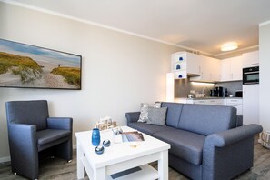 Ein modernes Wohnzimmer mit einem grauen Sofa, einem passenden Sessel und einem weißen Couchtisch. An der Wand hängt ein Gemälde, das eine Strandansicht zeigt.