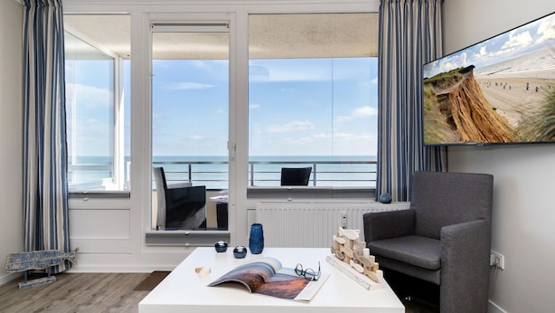Una habitación con una mesa blanca, un sillón gris y un ventanal con vista a la playa.