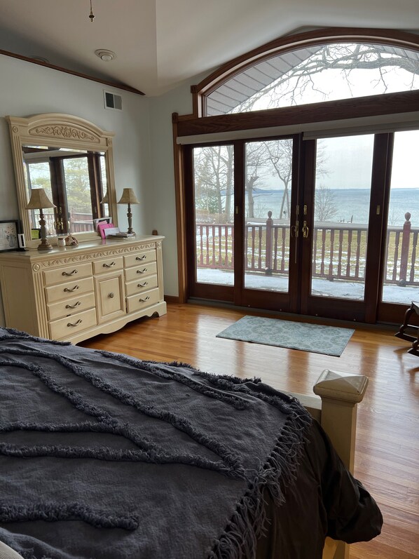 A bedroom with a large bed, a dresser, a mirror, and a view of a balcony with a railing and a body of water.