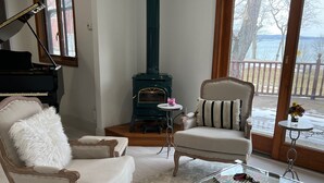 A living room with a piano, a fireplace, two armchairs, a glass coffee table, and a view of a deck with a railing. - Exclusive Lakefront Home Retreat w/Baby Grand Piano for Family FLX Fun & Friends (Ovid)