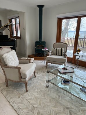 A living room with a piano, a fireplace, two armchairs, a glass coffee table, and a view of a deck with a railing.