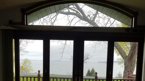 A room with a large sliding glass door leading to a deck with a view of a lake and a tree. - Exclusive Lakefront Home Retreat w/Baby Grand Piano for Family FLX Fun & Friends (Ovid)