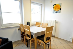Et spiseområde med et bord og stole, store vinduer, en radiator og et blomsterbillede på væggen.