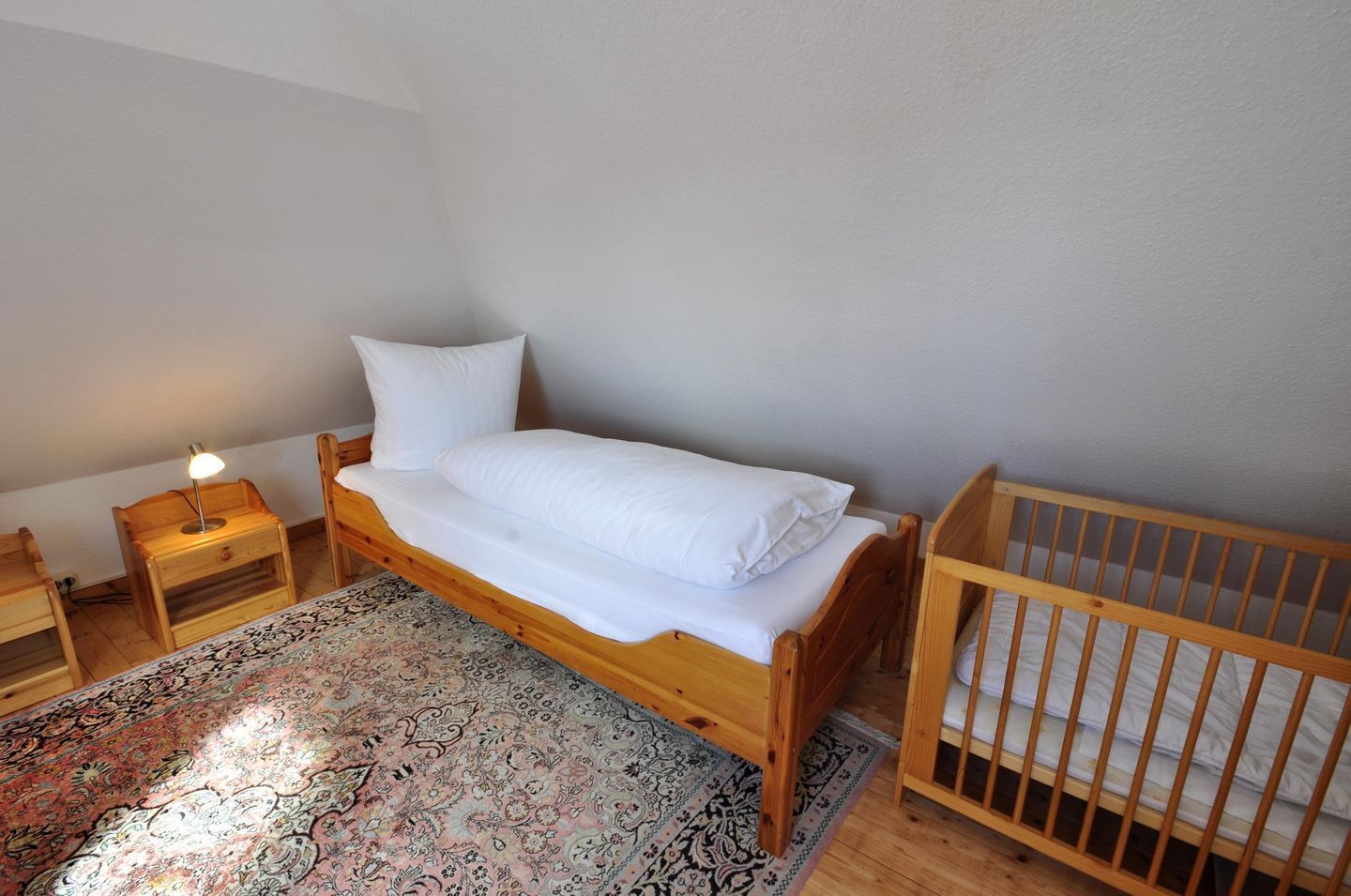 Een kleine, verzorgde slaapkamer met een enkel bed, een houten ledikant, een nachtkastje met een lamp en een kamerkleed met een patroon.