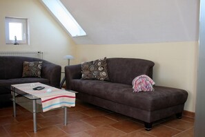 Uma sala de estar com um sofá marrom, uma mesa de centro de vidro e uma mesinha com um cobertor.