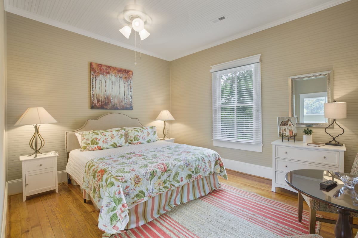 A bedroom with a bed, two bedside tables, a nightstand, a dresser, a chair, a table, a ceiling fan, a window with blinds, and a painting on the wall.