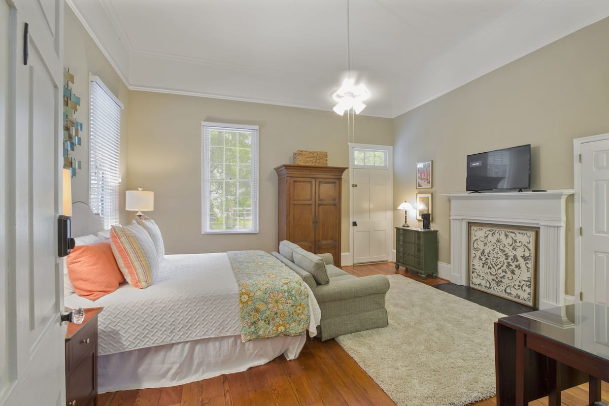 A bedroom with a bed, bedside table, lamp, dresser, television, fireplace, and a window with blinds.