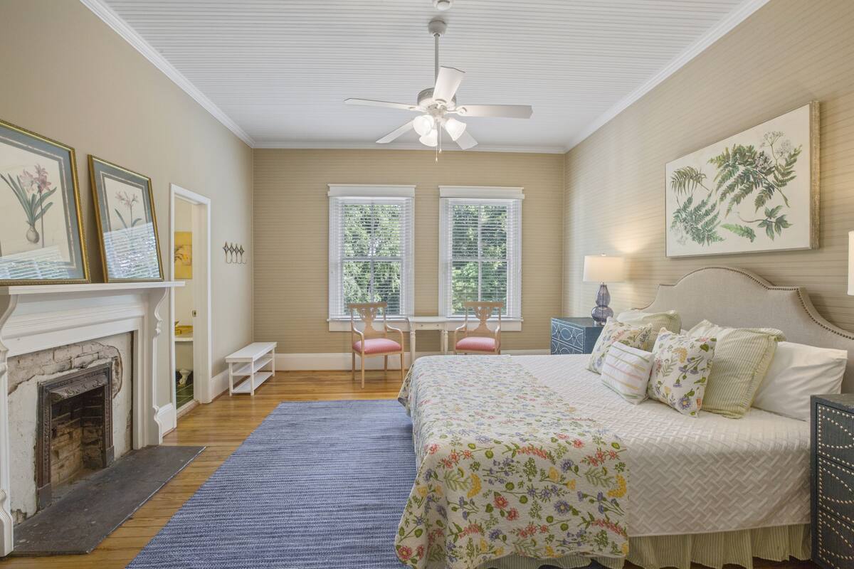 A bedroom with a bed, a fireplace, a ceiling fan, and two windows.