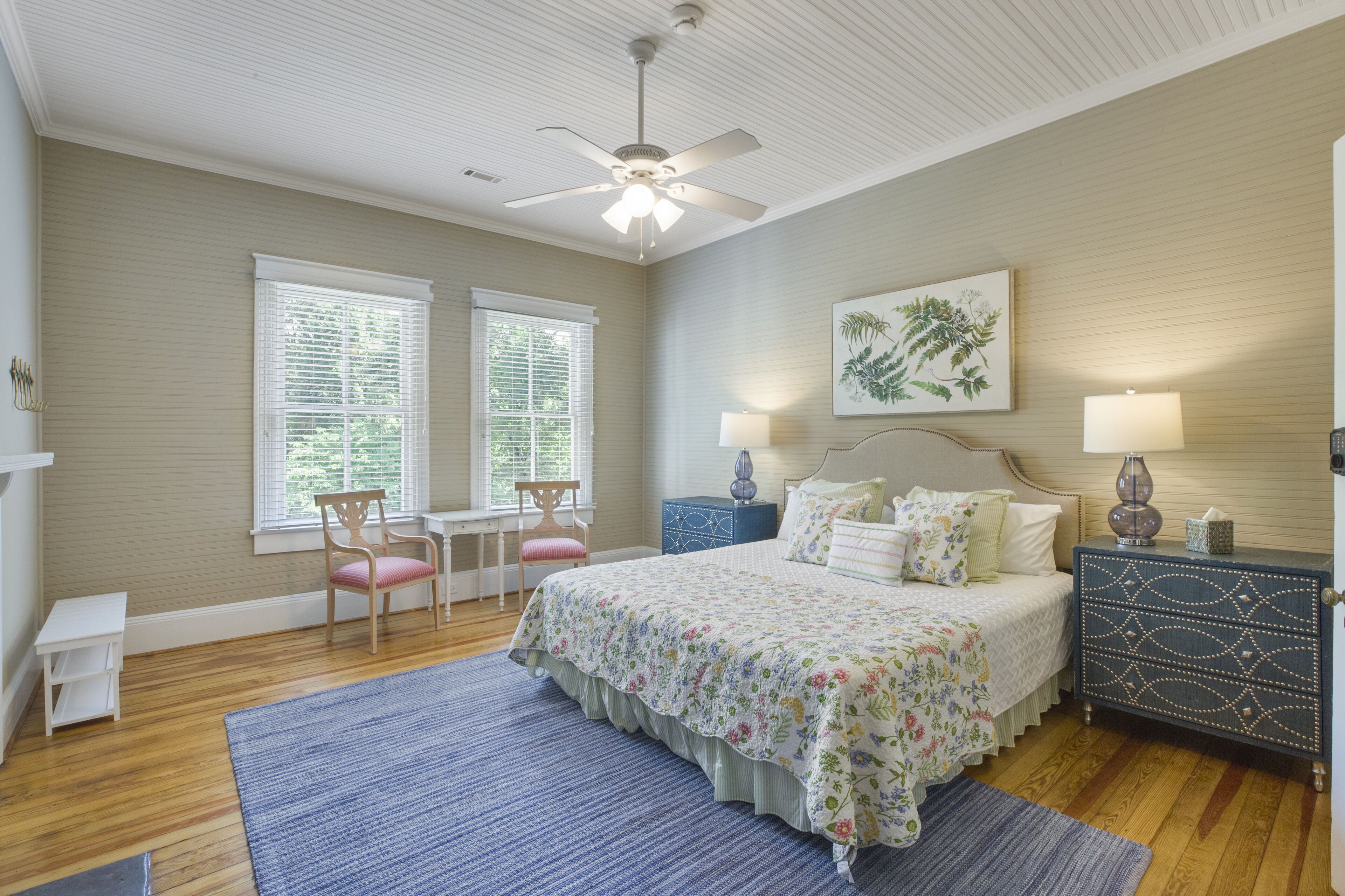 A bedroom with a bed, two bedside tables, a nightstand, a ceiling fan, and two windows with blinds.