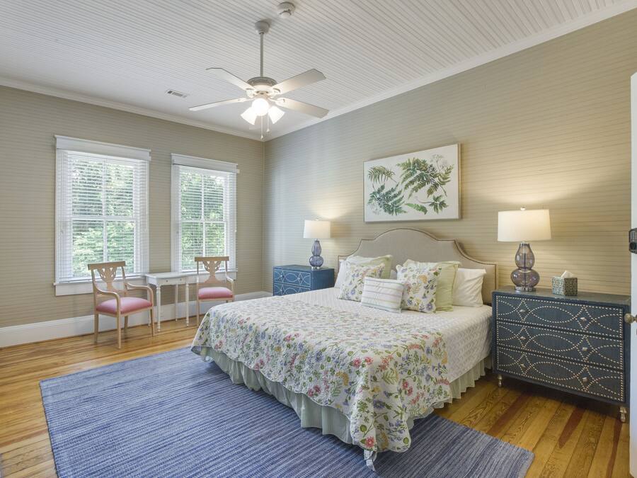 A bedroom with a bed, two bedside tables, a nightstand, a ceiling fan, and two windows with blinds.