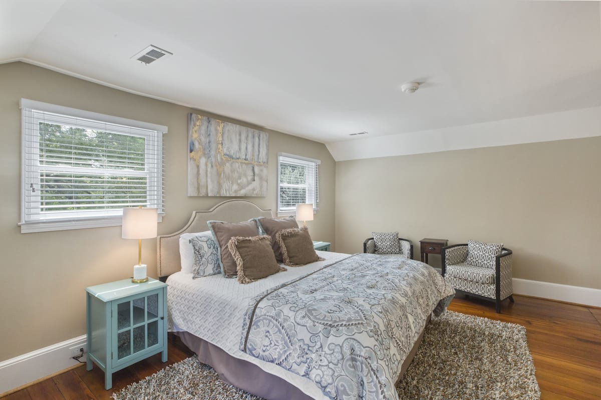 A bedroom with a large bed, two bedside tables, a chair, and a window with blinds.