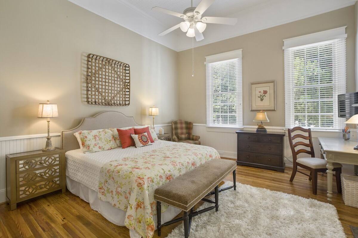 A bedroom with a bed, a desk, a chair, a bench, a ceiling fan, and a window with blinds.