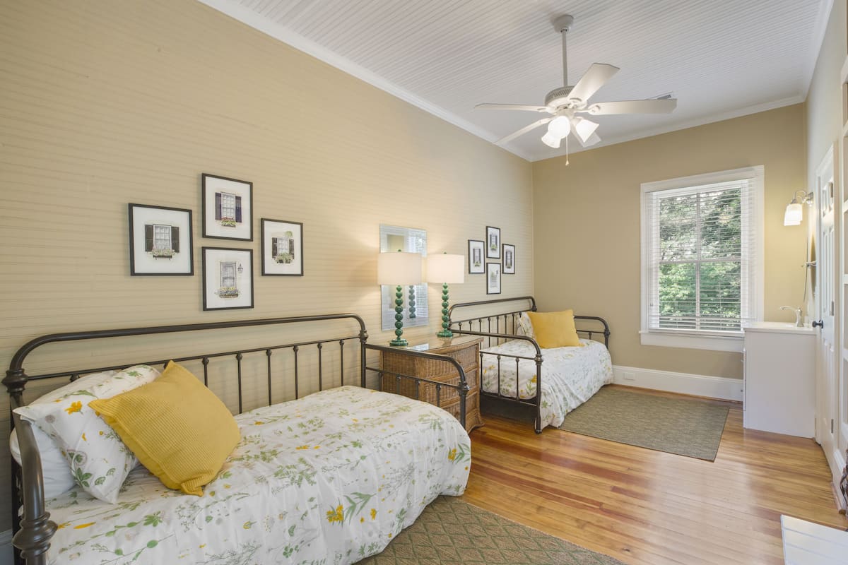 A bedroom with two beds, a ceiling fan, framed pictures on the wall, and a window with blinds.