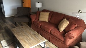 A living room with a wooden coffee table, a red sofa with two pillows, a grey armchair, a white floor lamp, and a wooden door. - Last minute opening for Cheyenne Frontier Days! (Cheyenne)