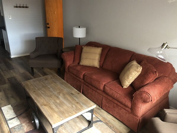 A living room with a wooden coffee table, a red sofa with two pillows, a grey armchair, a white floor lamp, and a wooden door.