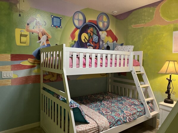 A bunk bed with a colorful mural on the wall, a small table with a lamp, and a switchboard.