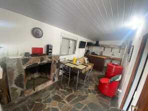 Una acogedora sala de estar con chimenea de piedra, una mesa de comedor con sillas, una zona de cocina con mostrador y electrodomésticos, y un reloj en la pared.