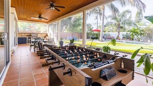 A spacious indoor area with a foosball table, a ceiling fan, and large windows offering a view of palm trees and a yard. - Luxury Resort Style, Heated Pool, 10 Mins to Gulf Mexico Beaches (Bradenton)