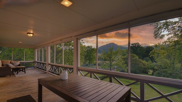 Une terrasse extérieure couverte, dotée d’une table et de chaises en bois, et de grandes fenêtres offrant une vue sur une forêt et des montagnes.