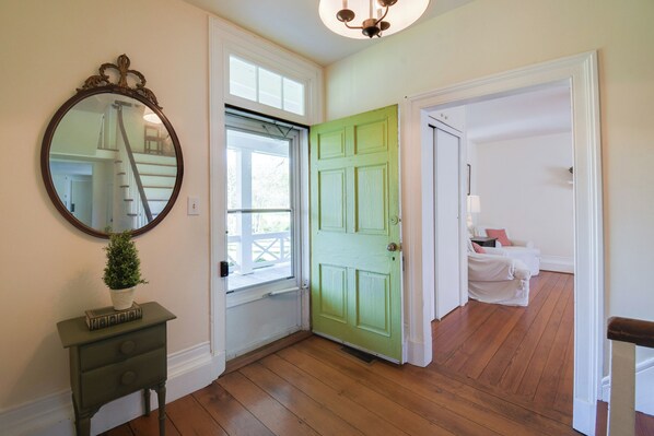 Una stanza con una porta verde, pavimento in legno, un tavolino con una pianta e uno specchio appeso al muro.