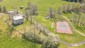 Vue aérienne d’une propriété rurale comprenant une grande maison, un petit abri de jardin, un court de tennis et de nombreux espaces verts.