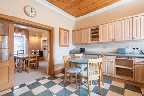 A kitchen with wooden cabinets, a dining table with chairs, a clock on the wall, and a checkered floor. - Scotston Villa - 5 bedroom house with large private back garden & hot tub (Forfar)
