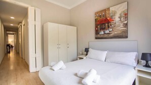 Habitación de hotel con una cama grande, un armario, un cuadro de una escena callejera y un pasillo que conduce a otra habitación.
