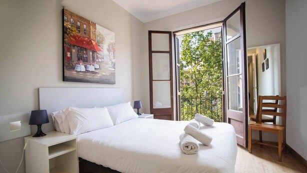 Habitación de hotel con cama, mesita de noche, una silla y ventana con vista a un balcón y árboles.