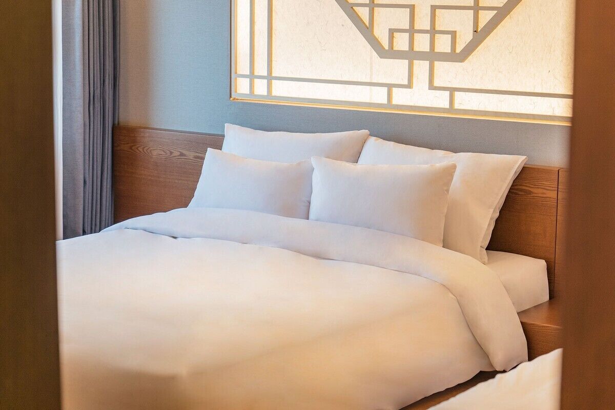 Cama bem arrumada com lençóis e almofadas brancos, cabeceira de madeira e uma obra de arte geométrica na parede acima.