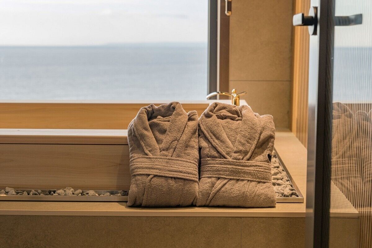 A window with a view of the sea, a wooden bench, and two towels placed on the bench.